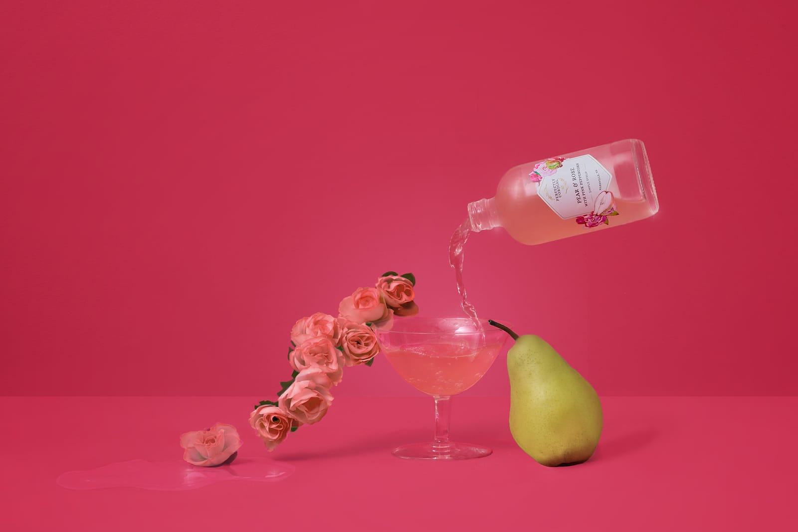 Pear Rose and Pink Peppercorn cordial pouring into a coupe glass beside a fresh pear on a rose pink backdrop