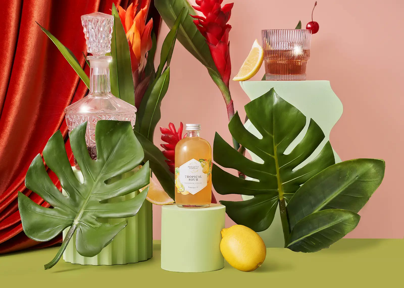 Tropical Sour cordial staged with tropical leaves, citrus, flowers, a decanter and a finished cocktail
